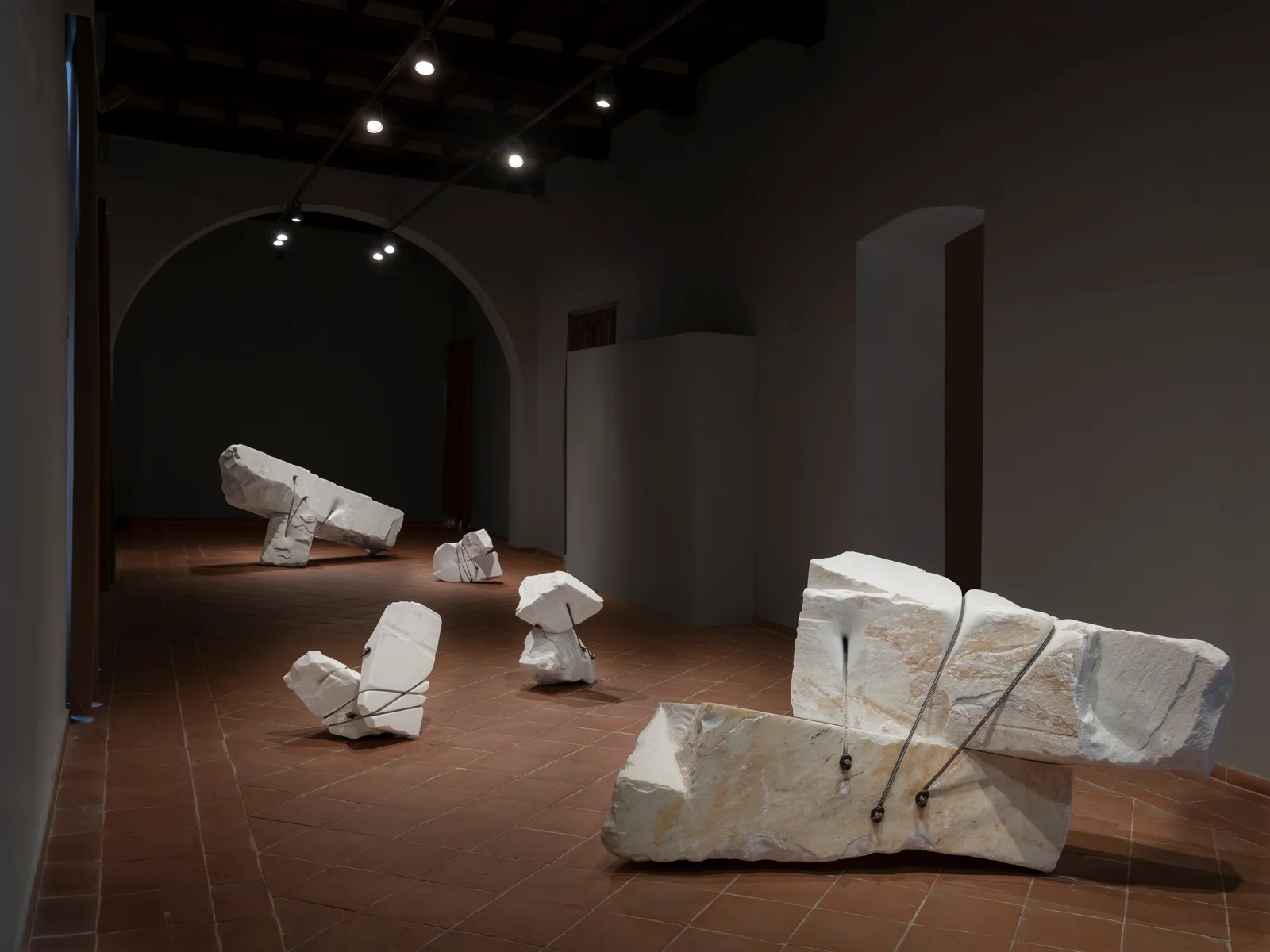White marble forms bound by steel cables, installed in a dark gallery at mudaC.